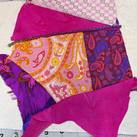 A crazy patch block being made from multiple patterned fabric panels in pink, purple, orange, and magenta, laid flat on a white sheet over a Fiskars cutting mat.
