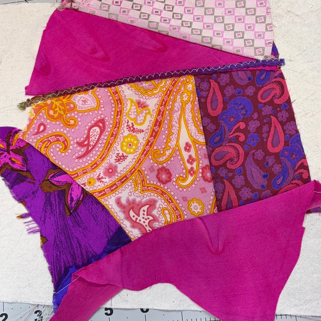 A crazy patch block being made from multiple patterned fabric panels in pink, purple, orange, and magenta, laid flat on a white sheet over a Fiskars cutting mat.