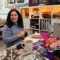 A woman with curly dark hair sits at a table in a sewing or craft studio, surrounded by colorful fabrics, shelves with sewing supplies, framed portraits on the wall, and a sewing machine. She is wearing a gray long-sleeve top.