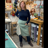 A woman stands in an art studio wearing a dark navy long-sleeve top, dark blue jeans, brown leather boots, and a sage green linen apron tied around her waist with a striped tea towel ucked in a loop.