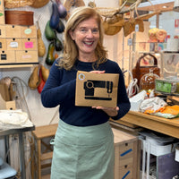 A smiling woman stands in a craft workshop holding a brown cardboard box with a black sewing machine graphic. She is wearing a navy long-sleeve top and a sage green apron.