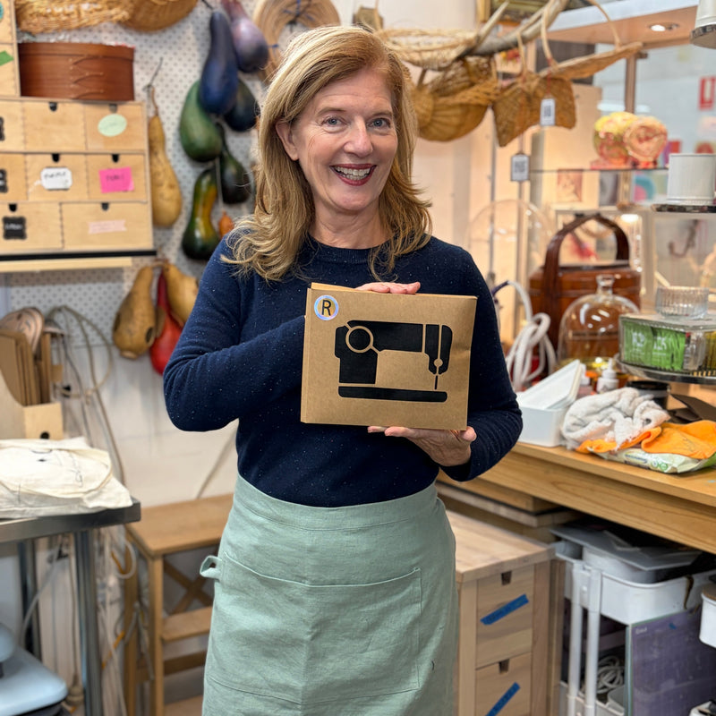 A smiling woman stands in a craft workshop holding a brown cardboard box with a black sewing machine graphic. She is wearing a navy long-sleeve top and a sage green apron.