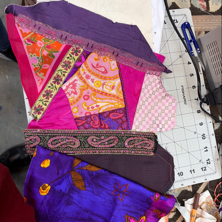 A pile of colorful patterned fabric pieces and a purple satin-like fabric piece with floral embroidery, arranged on a worktable with a cutting mat, scissors, and other sewing supplies visible.