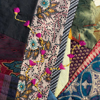A close-up of a patchwork quilt with multiple fabric panels, including a central cream patterned stripe with pink tassel embellishments, blue floral sections, and other multicolored textile patterns.
