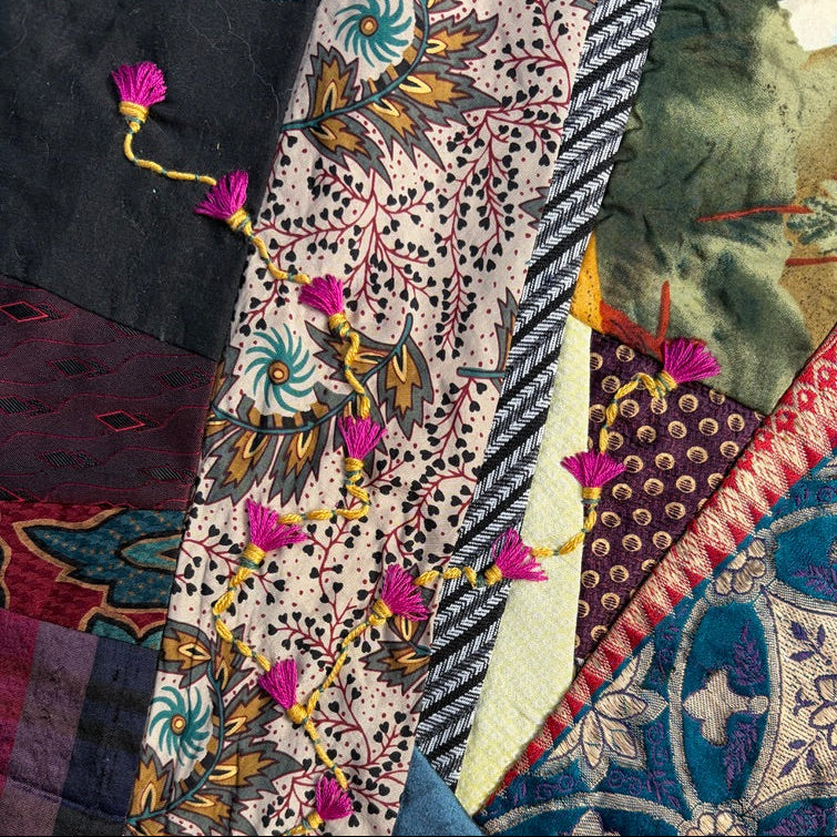 A close-up of a patchwork quilt with multiple fabric panels, including a central cream patterned stripe with pink tassel embellishments, blue floral sections, and other multicolored textile patterns.