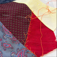 A close-up of fabric swatches laid out on a surface, including a navy textured piece, a cream woven section, a red stitched panel, a gray paisley textile with blue and red motifs, and a burgundy dotted fabric. A silver sewing needle rests on the materials.