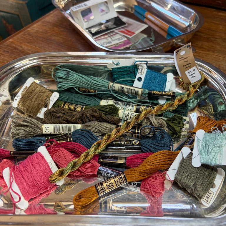 A collection of colorful embroidery floss skeins in various shades, including pink, green, blue, brown, and orange, arranged on a silver metal tray. One skein is braided and laid diagonally across the tray.