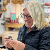 A woman with blonde hair and glasses, wearing a black quilted jacket, is shown in a workshop setting holding a small metal object in her hands.