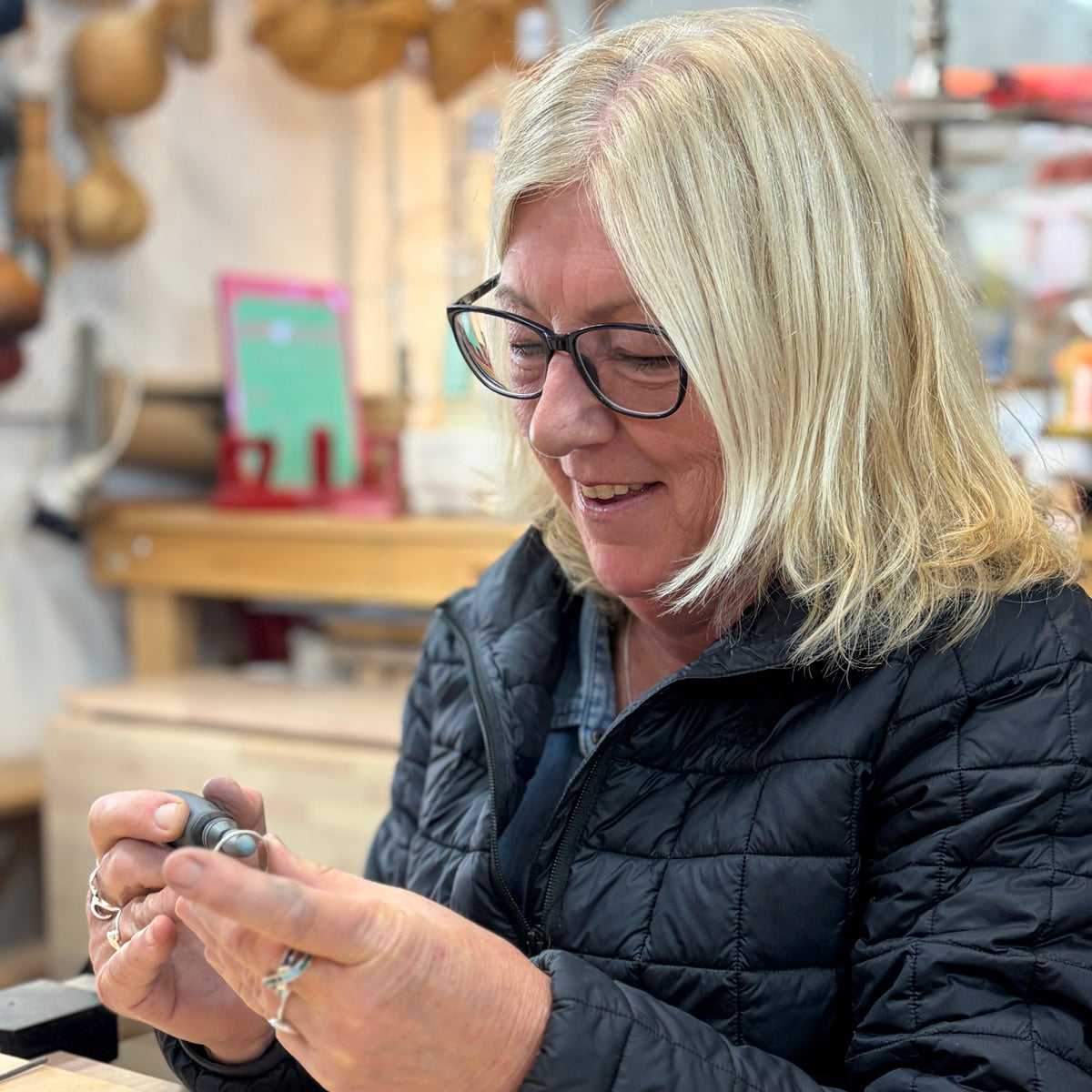 A woman with blonde hair and glasses, wearing a black quilted jacket, is shown in a workshop setting holding a small metal object in her hands.