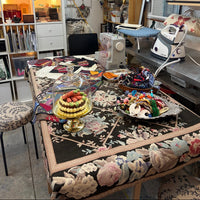 A highly detailed, cluttered craft studio featuring a prominent floral tablecloth on a table, alongside a sewing machine, iron, shelves of supplies, and decorative items.