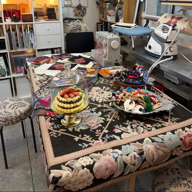 A highly detailed, cluttered craft studio featuring a prominent floral tablecloth on a table, alongside a sewing machine, iron, shelves of supplies, and decorative items.
