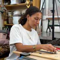 A woman with curly dark hair wearing a white t-shirt sits at a wooden workbench in a workshop, focused on crafting with metal tools and a hammer in the foreground.