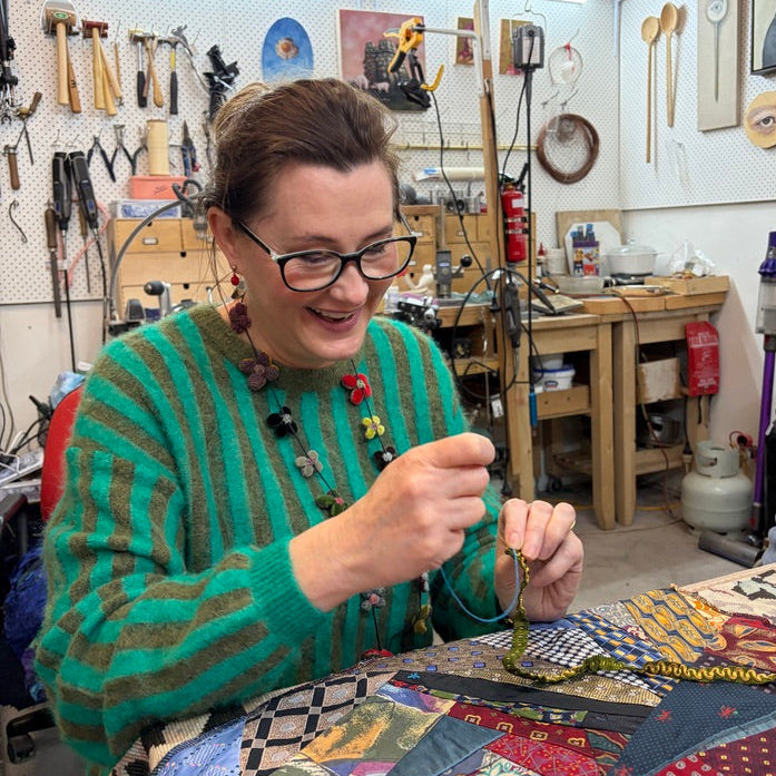 A woman with glasses and a green striped sweater sits at a table in a workshop, sewing a patchwork quilt made of colorful neckties. The background shows a cluttered but creative workspace filled with tools and art supplies.