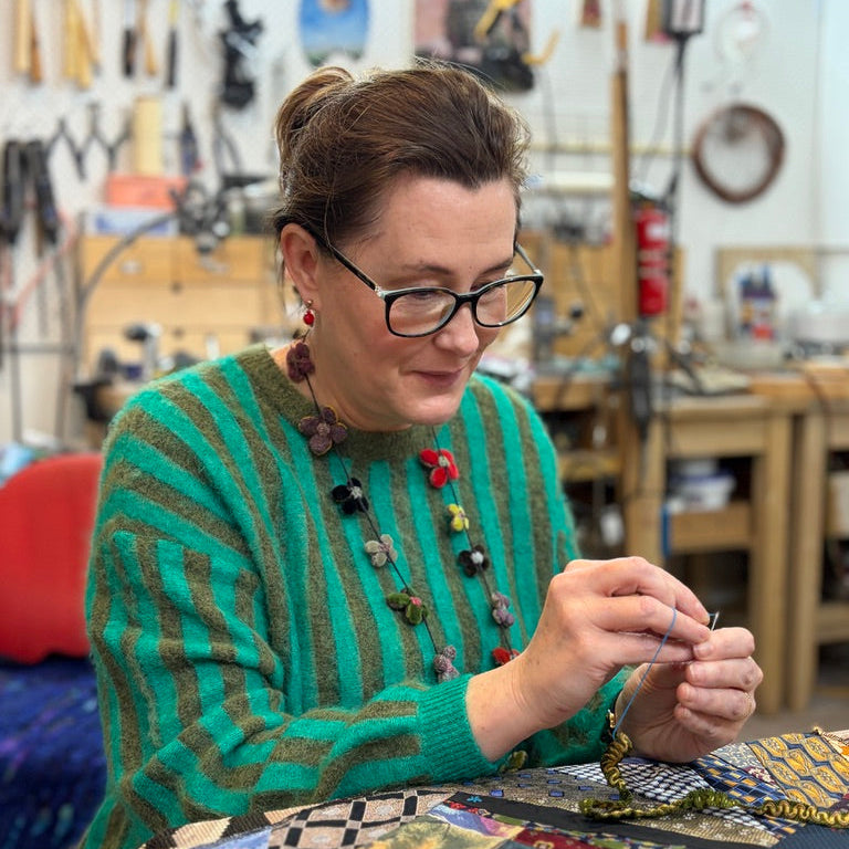 A woman wearing glasses and a green striped sweater sits at a table in a workshop, sewing a colorful patchwork quilt. Tools hang on the wall behind her, and she holds a needle and thread over the quilt.