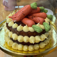 A small decorated cake under a clear glass cloche dome on a gold pedestal stand, set on a wooden table.