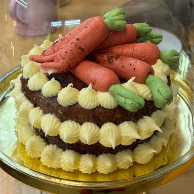 A small decorated cake under a clear glass cloche dome on a gold pedestal stand, set on a wooden table.