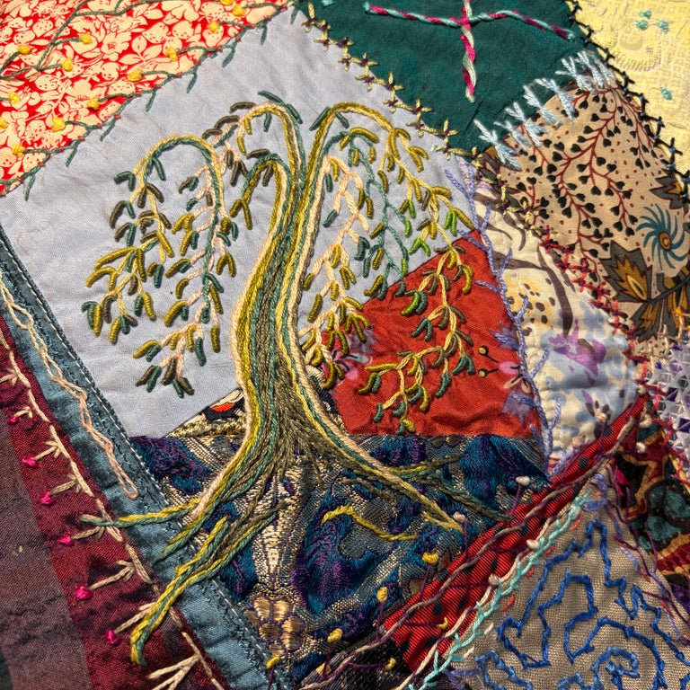 A close-up of a colorful patchwork quilt featuring a central embroidered tree motif with yellow-green branches and roots, surrounded by red, blue, green, and beige fabric sections with stitched patterns.