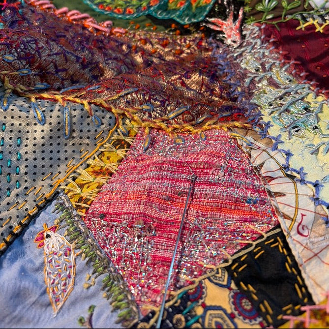 A close-up of a highly textured, multicolored patchwork quilt featuring intricate floral stitching, and various fabric panels in red, green, yellow, and blue.