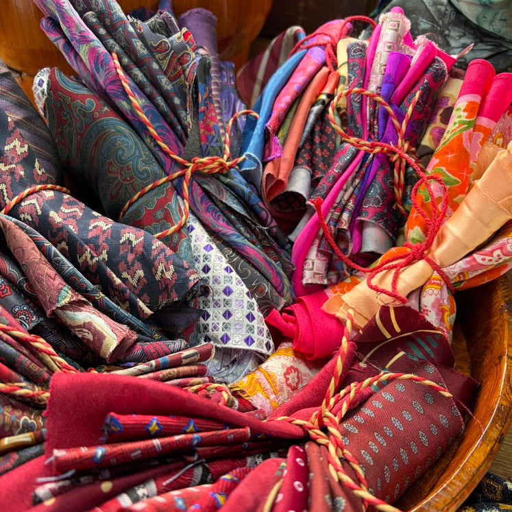 A close-up of multiple rolled and bundled pieces of patterned fabric tied with orange string, arranged in a wooden basket. The fabrics show varied designs including paisley, floral, polka dots, and geometric patterns in reds, pinks, blues, purples, and oranges.